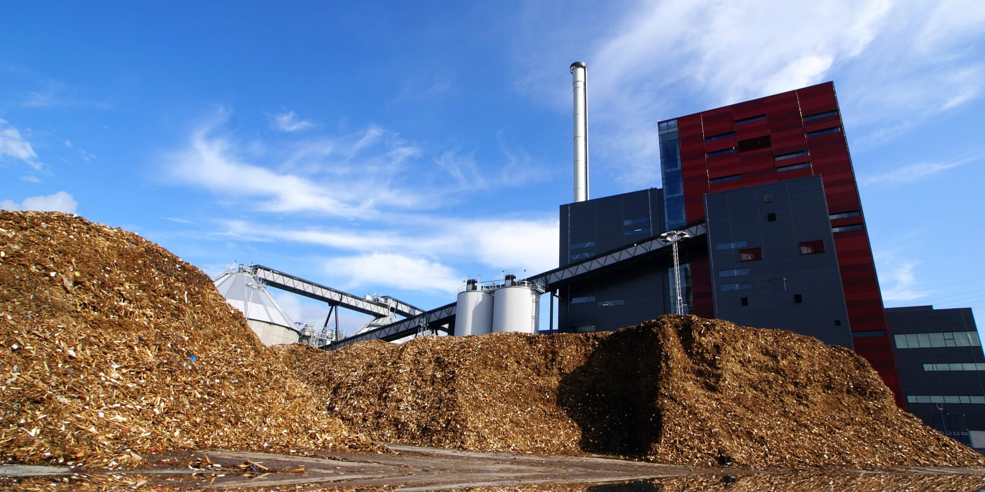 A bioenergy plant with a tall stack and conveyors above a large pile of wood chips